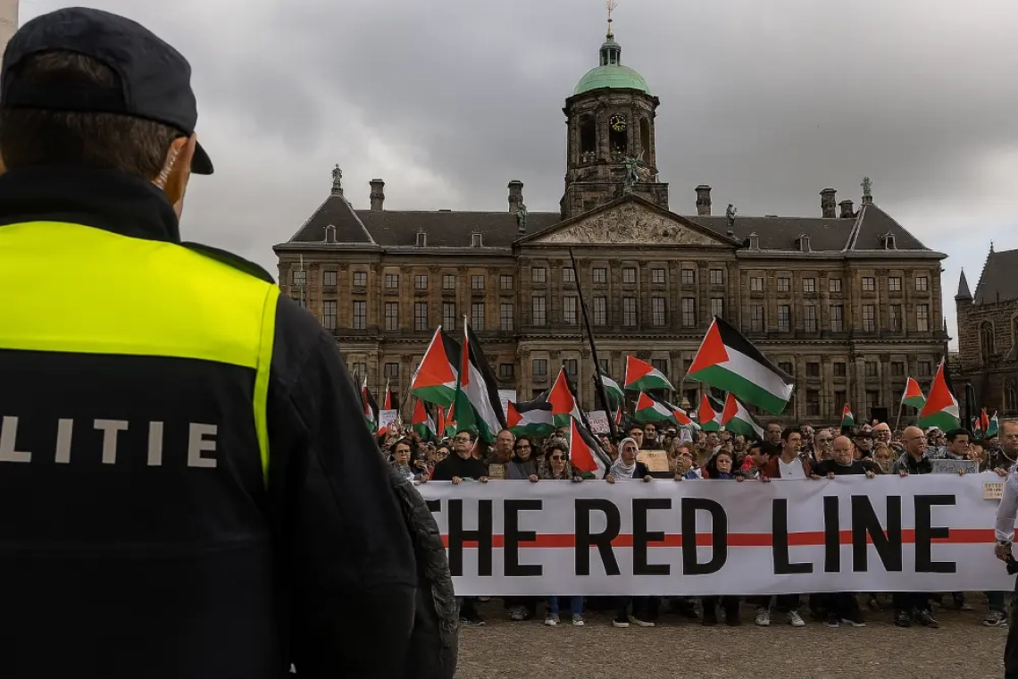 Nederlandse politieagent kijkt uit over een demonstratie op de Dam in Amsterdam, waar betogers met Palestijnse vlaggen en een groot protestspandoek zich verzamelen voor het Koninklijk Paleis.